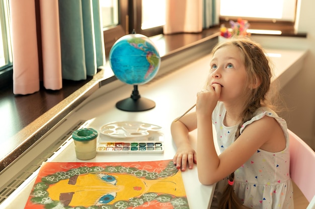 Retrato de menina está pintando com guache de cores e imagens de pincel enquanto está sentado à mesa no fundo da sala de casa