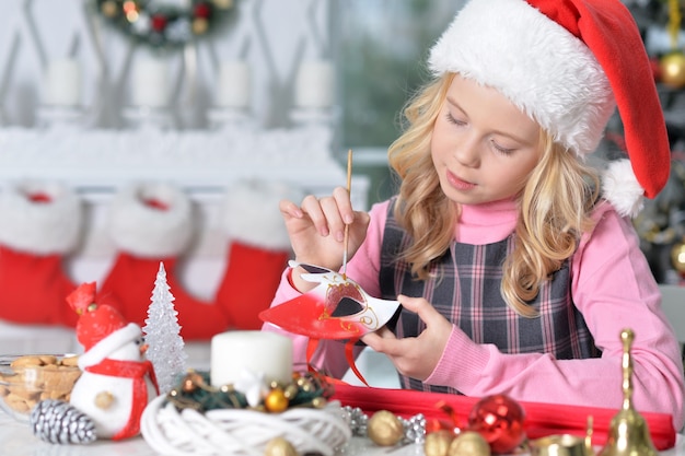 Retrato de menina feliz com chapéu de Papai Noel, se preparando para o Natal, sentado à mesa