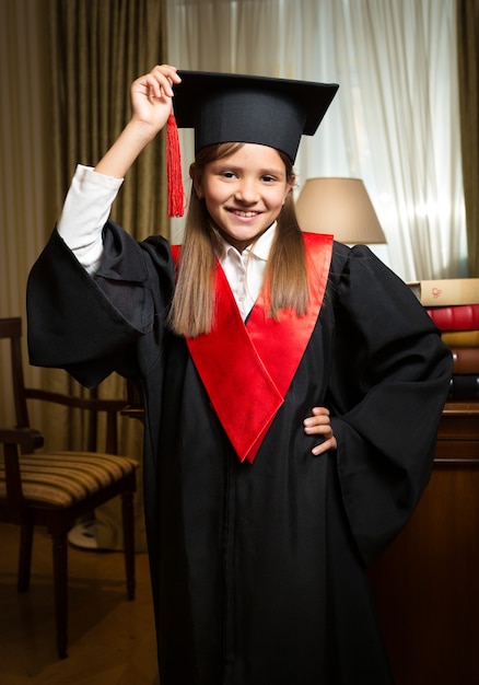 Retrato de menina feliz e sorridente com chapéu de formatura e vestido posando no interior clássico