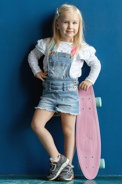 Retrato de menina feliz sorridente da criança com cabelos loiros, segurando um skate rosa enquanto posava no fundo da parede azul