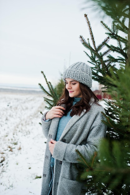 Retrato de menina gentil de casaco cinza e chapéu contra árvore de ano novo ao ar livre