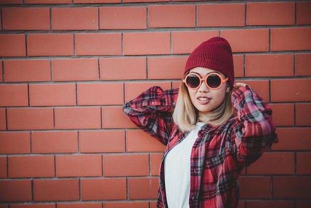 Retrato de menina hipster asiática no fundo da parede de tijolosela usa óculos de sol laranja e chapéu vermelho vintage estilo escuro conceito de moda