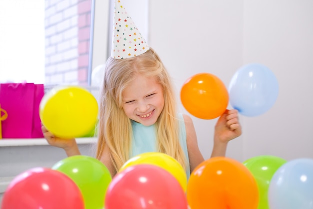 Retrato de menina loira caucasiana, sorrindo para a festa de aniversário de câmera. Fundo colorido festivo com balões. Foto vertical