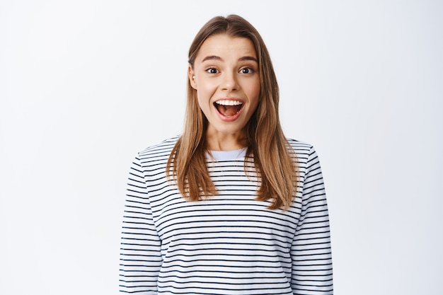 Retrato de menina loira surpresa e animada com a boca aberta fascinada, sorrindo maravilhada e olhando para algo legal, olhando o anúncio, parede branca