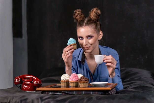 Retrato de menina modelo toma café da manhã na cama. Bandeja com cupcakes e café. Telefone retrô na cama.