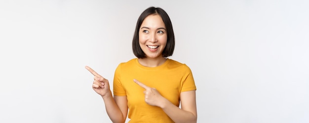 Retrato de menina morena asiática sorridente em camiseta amarela, apontando os dedos para a esquerda, mostrando o acordo promocional do espaço de cópia, demonstrando o banner em pé sobre fundo branco