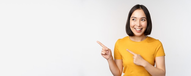 Retrato de menina morena asiática sorridente em camiseta amarela, apontando os dedos para a esquerda, mostrando o acordo promocional do espaço de cópia, demonstrando o banner em pé sobre fundo branco