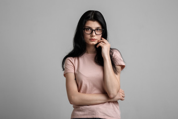 Retrato de menina morena linda inteligente em óculos com maquiagem natural, em fundo cinza.