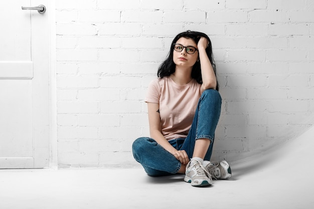 Retrato de menina morena linda inteligente em óculos com maquiagem natural, sentado perto da parede de tijolo branco.