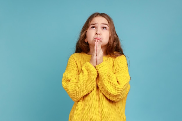 Retrato de menina olha para cima orando a Deus com olhos suplicantes fazendo desejo de infância e esperando vestindo suéter amarelo estilo casual Tiro de estúdio interior isolado em fundo azul