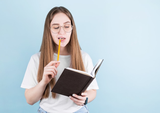 Retrato de menina pensativa pensativa, tendo o caderno e o lápis na boca. Isolado em azul com espaço de cópia.