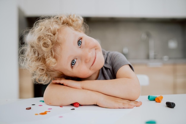 Retrato de menina pré-escolar sorridente, sentada à mesa da cozinha, brincando com plasticina