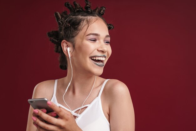 Retrato de menina punk feliz com penteado bizarro e batom escuro rindo enquanto usa o smartphone com fones de ouvido isolados sobre a parede vermelha