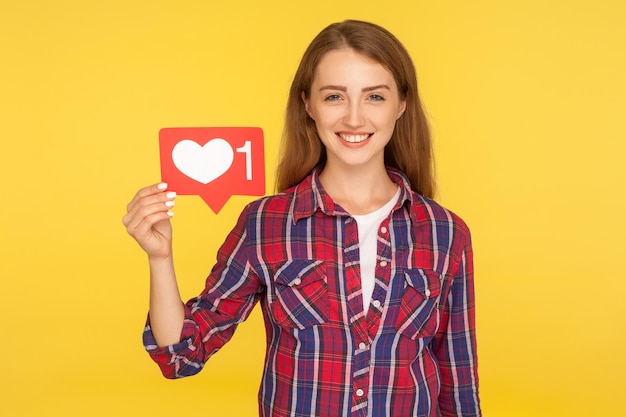 Foto retrato de menina ruiva alegre em camisa quadriculada sorrindo e segurando mídias sociais como botão de coração de ícone para desfrutar de conteúdo inscreva-se e siga tiro de estúdio interno isolado em fundo amarelo