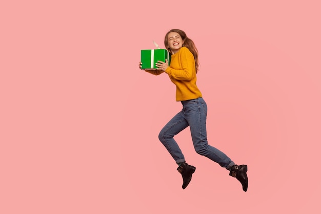 Retrato de menina ruiva extremamente feliz na camisola e jeans pulando no ar com caixa de presente, cheia de energia e entusiasmo voando com presente de férias. tiro de estúdio interior isolado no fundo rosa