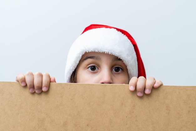 Retrato de menina sorridente com chapéu de Natal. Garoto espiando atrás do tabuleiro de férias em branco. Criança feliz e fofa posando atrás do painel de papel kraft, isolado no fundo branco.