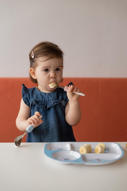 Retrato de menina tem a primeira alimentação segurando banana no garfo em casa Criança muito estilosa