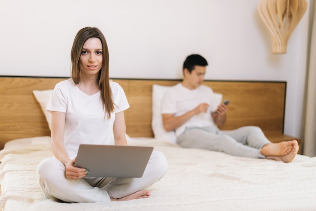 Retrato de menina trabalhando no laptop na cama. Guy usa o telefone atrás da namorada. Jovem olhando para a câmera. A menina está digitando no bloco de notas, o cara está navegando na Internet