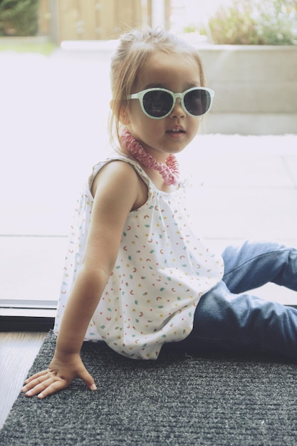 Foto retrato de menina usando óculos de sol enquanto está sentada contra a janela em casa