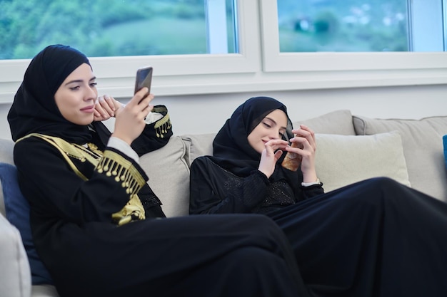 Retrato de meninas árabes usando abaya moderno usando telefone celular. Mulheres muçulmanas com hijab sentadas no sofá representando tecnologia