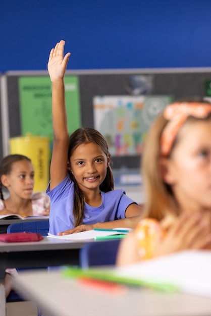 Foto retrato de meninas de escola felizes e diversas em mesas levantando as mãos na sala de aula