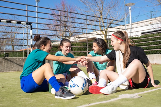 Retrato de meninas passando fins de semana no campo de futebol. Quatro garotas sorridentes em roupas esportivas sentadas no chão, descansando de mãos dadas conversando sobre o jogo. Estilo de vida saudável, conceito de esporte de equipe