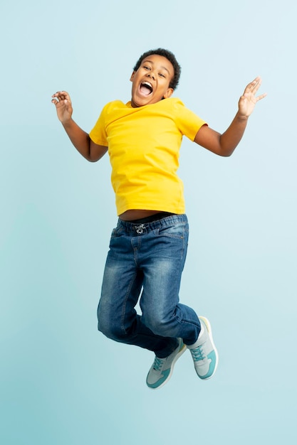 Retrato de menino afro-americano positivo sorridente vestindo roupas casuais elegantes pulando se divertindo olhando para a câmera isolada no fundo azul conceito de infância