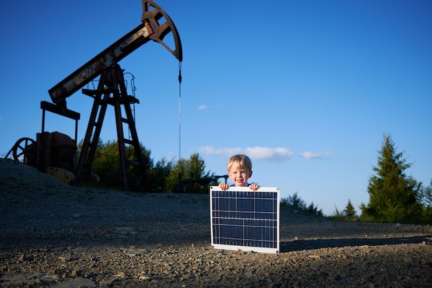 Retrato de menino alegre com bateria solar sorrindo para a câmera durante o tempo de educação na estação de petróleo com torre de bomba no fundo criança feliz com painel para energia verde alternativa de produto