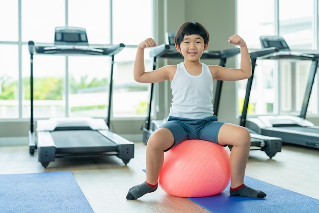 Retrato de menino asiático mostrando músculos sentados na bola de ginástica após exercícios no clube esportivo de fitness