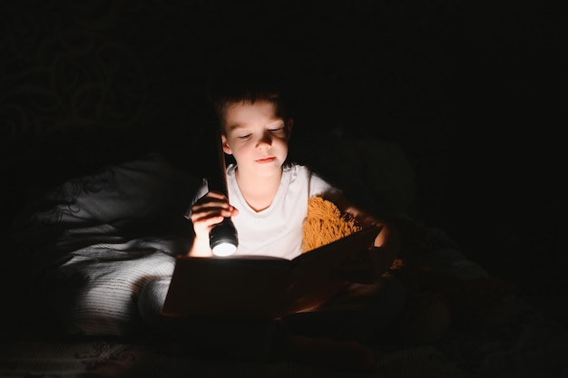 Retrato de menino bonitinho lendo na cama com lanterna no quarto escuro curtindo contos de fadas