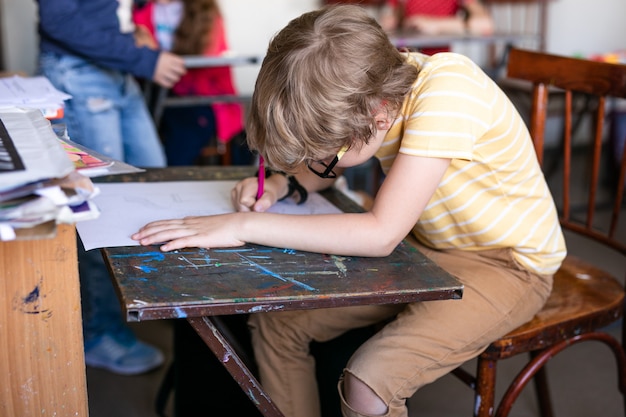 Retrato de menino bonito com caneta de desenho e papel na mesa na sala de aula.