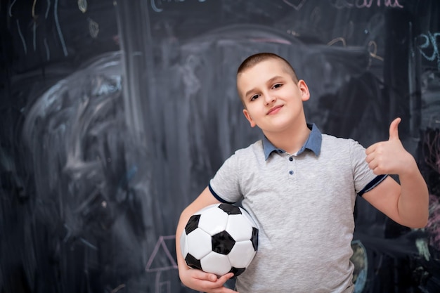 retrato de menino bonito feliz se divertindo segurando uma bola de futebol em pé na frente do quadro negro