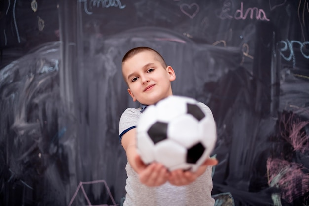 retrato de menino bonito feliz se divertindo segurando uma bola de futebol em pé na frente do quadro negro