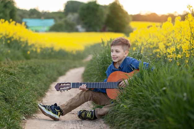 Retrato de menino bonito tocando uma guitarra sentado na estrada no campo amarelo de verão.