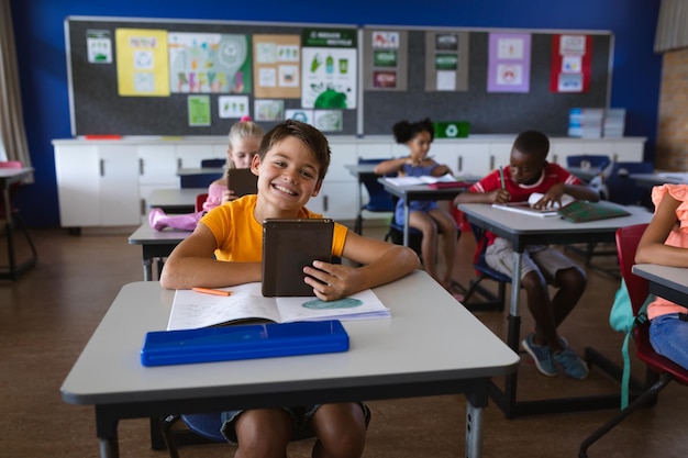 Retrato de menino caucasiano sorridente segurando tablet digital sentado na mesa na escola primária