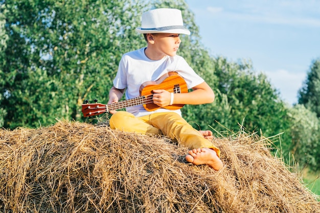 Retrato de menino descalço no chapéu no palheiro no campo Tocando ukulele de guitarra pequeno Dia ensolarado Vista lateral ao ar livre