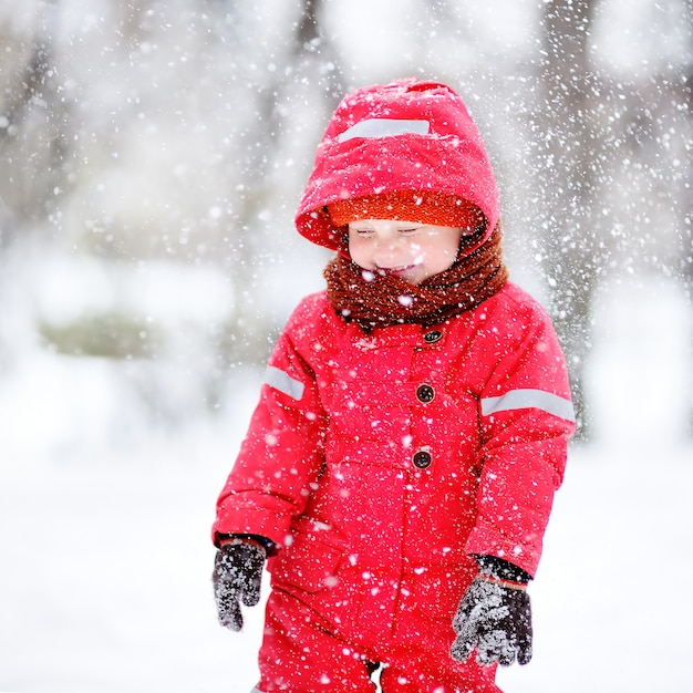 Retrato de menino em roupas de inverno vermelho se divertindo com neve durante a queda de neve