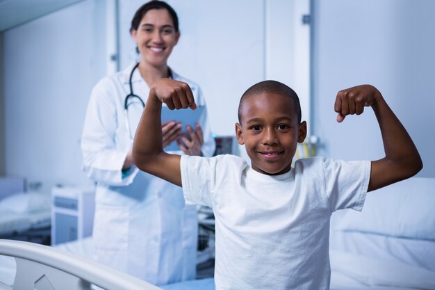 Retrato de menino flexionando seus músculos e médica em pé no fundo