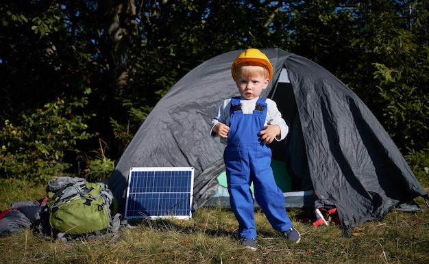 Retrato de menino focado no capacete de construção em pé na grama em frente à tenda um painel solar e mochila para caminhada Geração jovem demonstrando nova ecoenergia solar