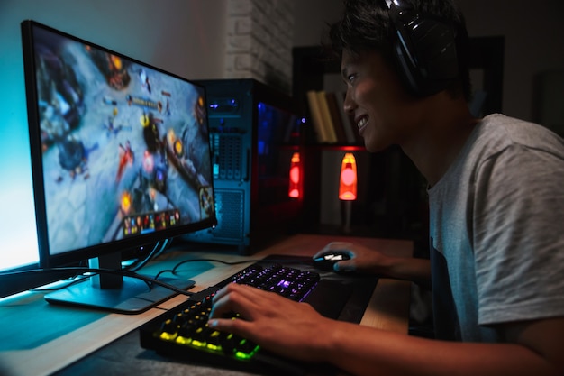 Retrato de menino jogador asiático de 16-18 anos, sorridente, jogando videogame no computador em um quarto escuro, usando fones de ouvido e um teclado colorido com luz de fundo