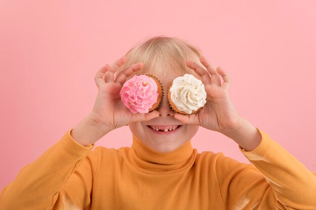 Retrato de menino loiro fecha os olhos com dois cupcakes com creme rosa e branco sobre fundo rosa. As crianças adoram doces.