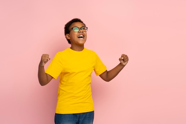 Retrato de menino nigeriano sorridente animado usando óculos e elegante camiseta amarela e jeans fazendo gesto vencedor olhando para longe isolado no fundo rosa cópia espaço conceito de vitória