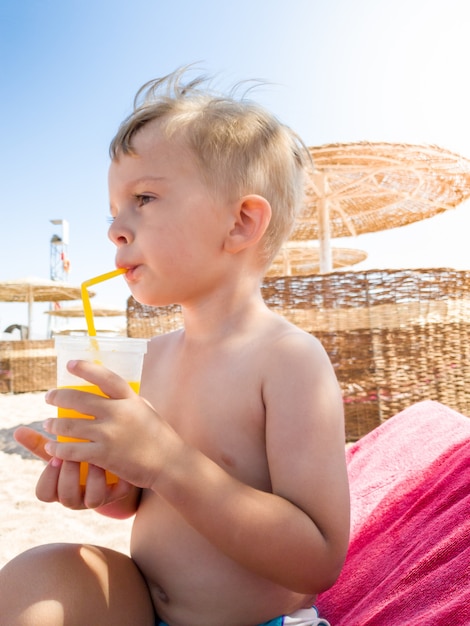 Retrato de menino sentado na praia do mar e bebendo suco de laranja da palha. Crianças relaxando e se divertindo durante as férias de verão.