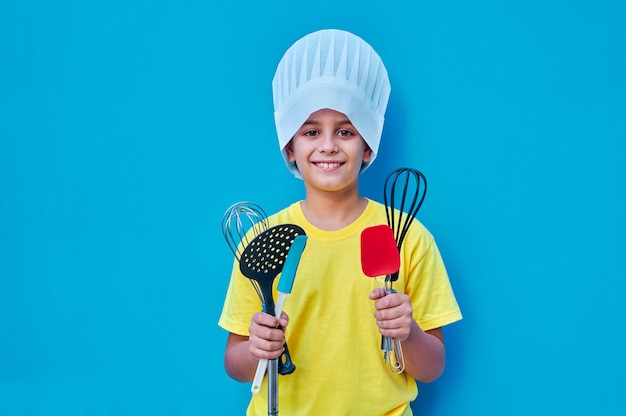 Retrato de menino sorridente com camiseta amarela e chapéu de chef, com utensílios de cozinha prontos para aprender a cozinhar