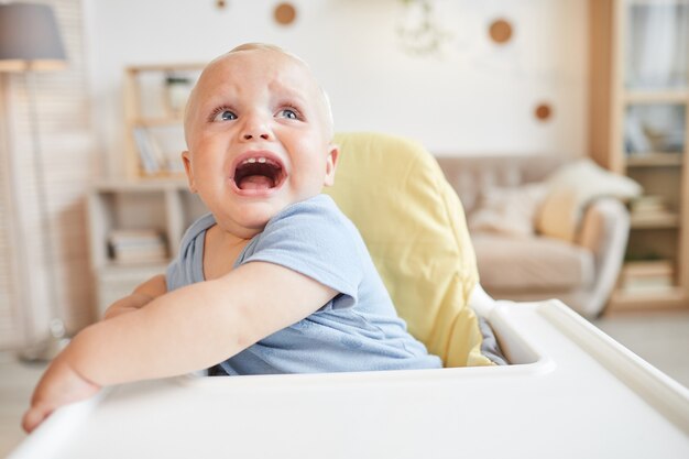 Retrato de menino travesso sentado em uma cadeira alta chorando por alguma coisa