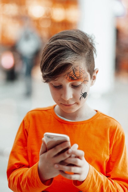 Retrato de menino zoomer autêntico sincero com pintura de rosto usando telefone celular na festa de Halloween