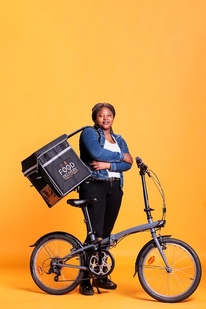 Retrato de mensageiro sorridente em pé ao lado da bicicleta segurando a mochila térmica de entrega, entregando o pedido de almoço ao cliente. Trabalhador de pizzaria em pé no estúdio sobre fundo amarelo