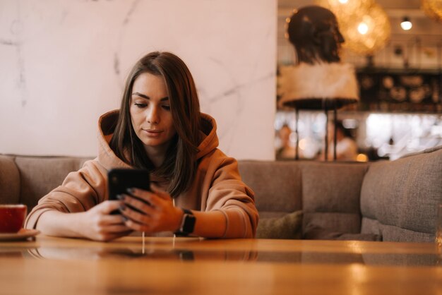 Retrato de mensagem de digitação de mulher jovem e atraente no telefone no café aconchegante. Menina bonita em roupas casuais pensa enquanto digita no telefone no café. Conceito de atividade de lazer.