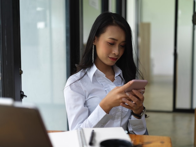 Retrato de mensagens de texto femininas no smartphone enquanto está sentado na mesa de trabalho na sala de escritório