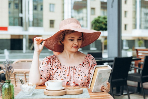 Retrato de moda ao ar livre de uma mulher deslumbrante sentada em um café eu bebo café e leio um livro antigo uma mulher de vestido e chapéu